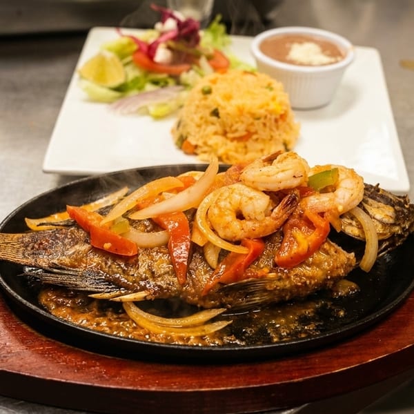 Steaming whole fish with shrimp, peppers, and onions in a skillet, with rice and beans.